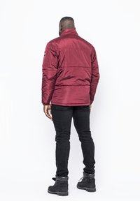 Giacca trapuntata borgogna con colletto alto, caratterizzata da dettagli di cucitura orizzontale. Indossata sopra jeans neri e stivali. Vista da dietro.