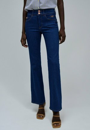 Frau trägt hochtaillierte, dunkelblaue Schlagjeans mit Kupferknöpfen und hellbraunen Riemchensandalen, steht vor einem schlichten hellen Hintergrund.