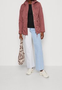 Růžová bunda s kapucí a předním zipem, černý top, bílé džíny s širokými nohavicemi a tenisky. Drží hnědo-krémovou vzorovanou tašku.