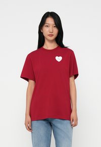 O tânără cu păr lung, negru, poartă un tricou roșu cu o inimă albă mică și textul "Hugo", asortat cu blugi bleu deschis.