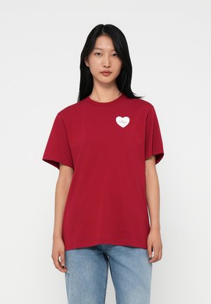 Giovane donna con lunghi capelli neri che indossa una maglietta rossa con un piccolo cuore bianco e il testo "Hugo", abbinata a jeans azzurri chiari.