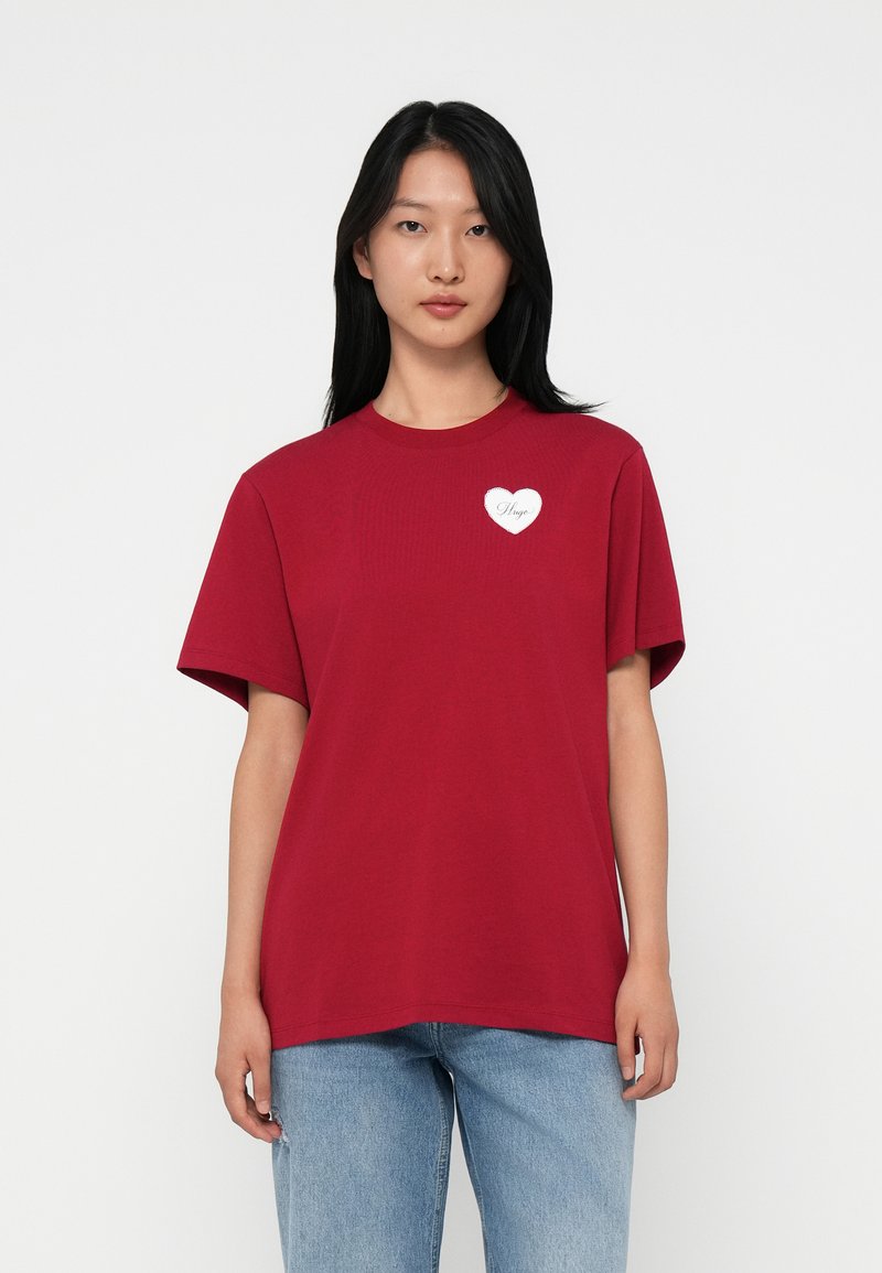 Joven con cabello largo y negro que lleva una camiseta roja con un pequeño corazón blanco y el texto "Hugo", combinada con jeans azul claro.