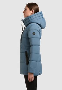 Hele sinine puffer-jakk suure volditud kapuutsiga, külgpocketitega ja elastsete mansettidega. Sellel on must logotäht sleeve'il.