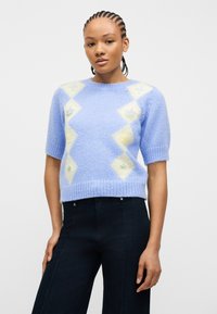 Γυναίκα με πλεξούδες που φοράει ανοιχτό μπλε χνουδωτό πουλόβερ με κοντά μανίκια και διαμάντι σχέδια, μαζί με σκουρόχρωμο ψηλόμεσο παντελόνι, σε λευκό φόντο.