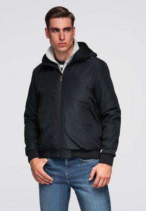 Jeune homme portant une veste à capuche noire, une couche intérieure grise et un jean bleu, posant avec les mains près des poches sur un fond uni.