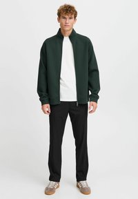 Dark green zip-up jacket with a high collar, paired with a white shirt and black trousers. Features relaxed fit and smooth texture.