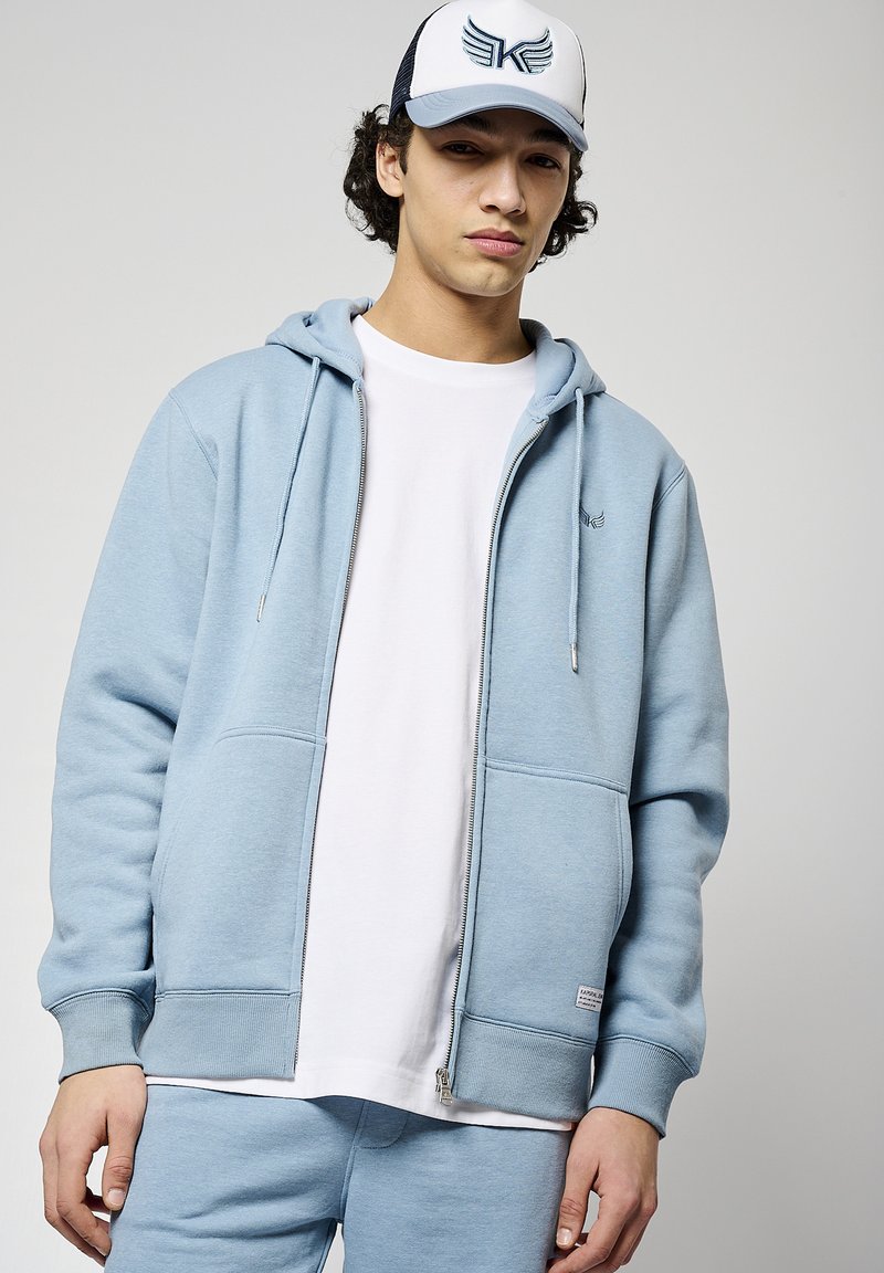 Sudadera con cremallera de color azul claro, con dos bolsillos frontales, textura suave y cordones, combinada con una camiseta blanca y pantalones de chándal a juego.
