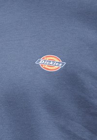 Μικρό λογότυπο Dickies με λευκό κείμενο σε μπλε ορθογώνιο φόντο, τοποθετημένο πάνω σε στρογγυλό πορτοκαλί και κίτρινο εικονίδιο, τυπωμένο σε μπλε ύφασμα.