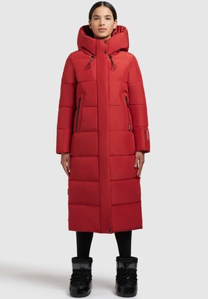 Vrouw in een lange, rode, gewatteerde winterjas met een hoge kraag, twee ritszakken en zwarte winterlaarzen op een neutrale achtergrond.