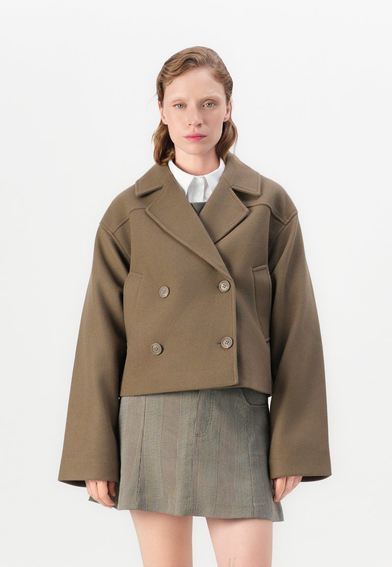 Cropped, double-breasted jacket in olive green wool blend with large buttons and wide sleeves, paired with a gray plaid skirt.