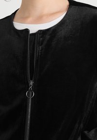 Svart sammet bomberjacka med rund halsringning, framficksdragkedja och slät textur, med en subtil glans.