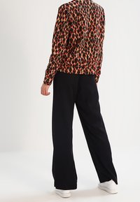 Personne portant un haut à manches longues rouge et noir à imprimé léopard, un pantalon large noir et des baskets blanches, debout devant un fond uni.