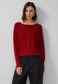 Roter gestrickter Pullover mit ribbeligem Saum und weitem Halsausschnitt, ausgestattet mit langen Ärmeln und einer weichen Textur. Kombiniert mit schwarzen Hosen.