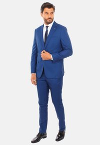 Suit blu su misura con taglio slim, pantaloni abbinati, giacca monopetto e revers a notch. Scarpe nere lucide completano il look.