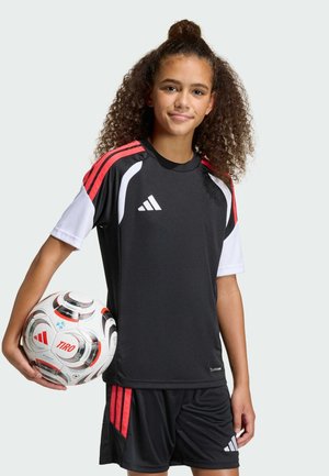 Dívka s kudrnatými vlasy držící fotbalový míč, oblečená v černo-bílém sportovním oblečení Adidas s červenými pruhy, stojící před jednobarevným pozadím.