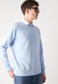 Jeune homme portant des lunettes de soleil noires, un sweat-shirt bleu clair par-dessus une chemise blanche à col, et un jean bleu, debout devant un fond blanc.