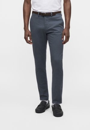 Homme portant un pantalon gris slim, des chaussettes blanches, des mocassins noirs, une chemise blanche rentrée dans le pantalon et une ceinture marron, devant un fond uni.