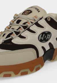 Baskets beige et noires avec semelle marron, avec le logo DC sur le côté et la languette, lacets beiges passant dans des boucles avec des accents orange.