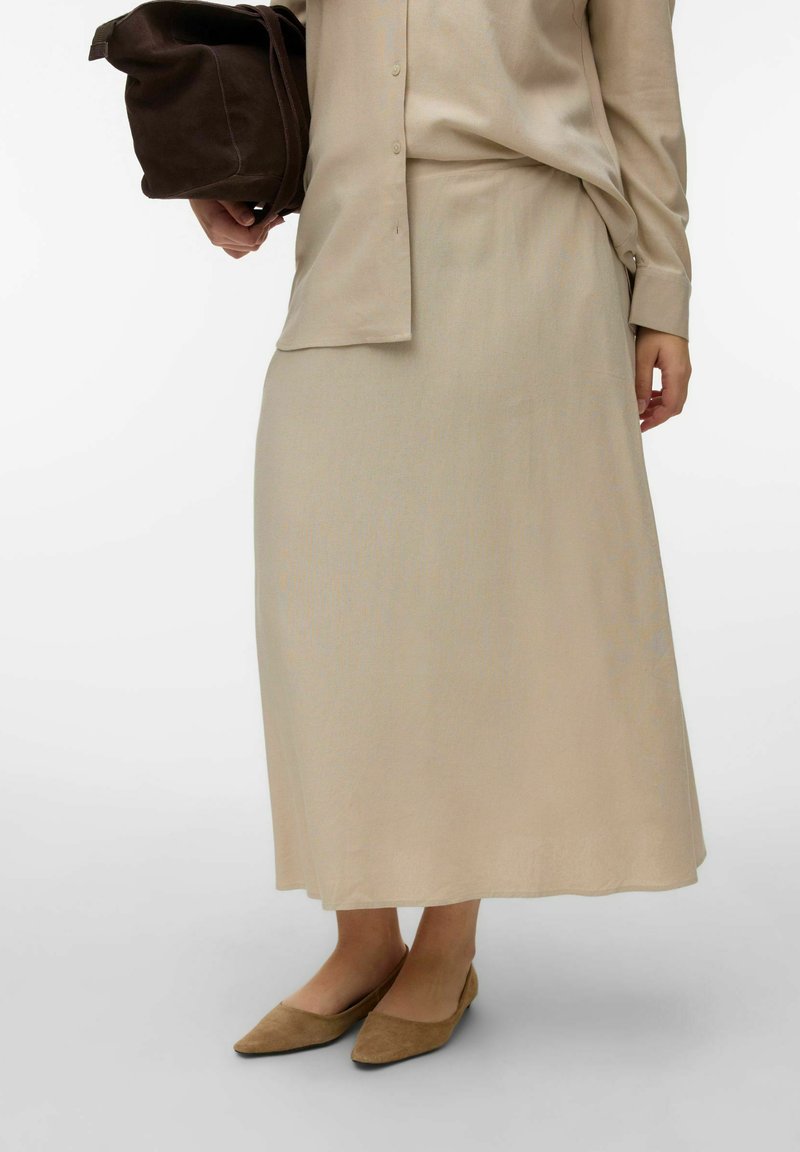 Femme portant une longue jupe beige et une chemise boutonnée, tenant un sac à main marron foncé et chaussée de ballerines pointues couleur fauve.