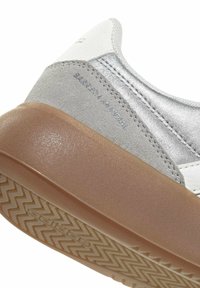 Chaussure basse en daim gris et argent métallique avec des accents blancs et une semelle en caoutchouc. Surface texturée notable et motif de bande de roulement distinctif.