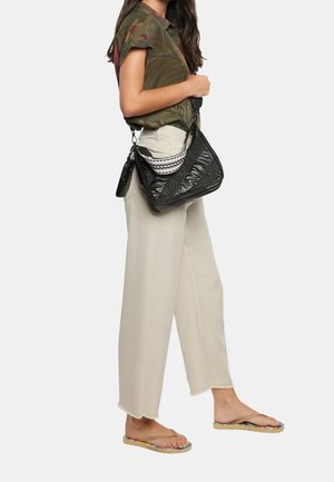 Mujer vistiendo pantalones anchos beige, camisa de manga corta con estampado floral verde, sandalias tipo flip-flop y llevando un bolso de hombro negro texturizado con un pequeño monedero.