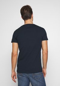 Tmavě modré bavlněné tričko s krátkým rukávem, kulatým výstřihem a slim fit, kombinované se světlými modrými džínami. Jednoduchá, hladká textura.