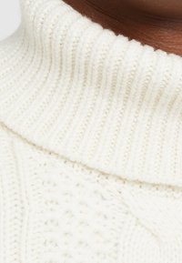 Maglione bianco lavorato a maglia con un alto colletto a coste e motivi testurizzati. Il tessuto è morbido e spesso, evidenziando il design stratificato della maglia.