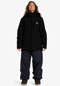 Chaqueta negra impermeable con cierre de cremallera, capucha ajustable y bolsillos frontales, combinada con pantalones negros de corte holgado y zapatos negros.