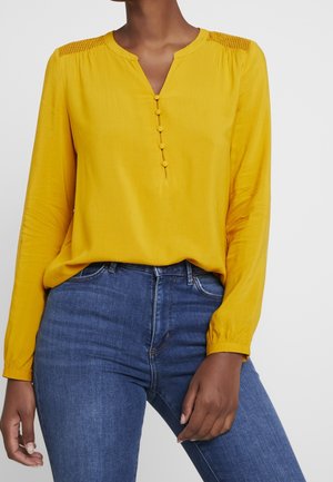 Kobieta w żółtej bluzce z długim rękawem, z guzikami i teksturowanymi ramionami, zestawiona z wysokim stanem niebieskich dżinsów.
