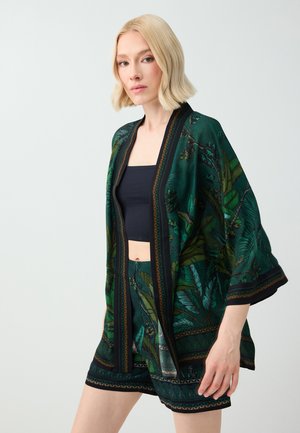 Woman with short blonde hair wears a green patterned kimono-style jacket and matching shorts over a black crop top against a plain background.