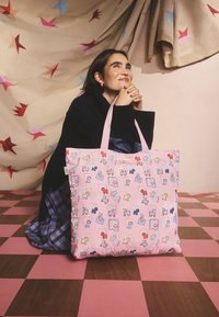 Jauna moteris tamsiu megztiniu ir languota sijonu klūpo ant rožinės ir rudos languotos grindų dangos, laiko didelę rožinę rankinę su spalvingu šuns raštu.