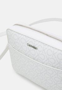 Calvin Klein hvid crossbody taske lavet af struktureret materiale, med en sølv lynlås, subtilt logo mønster og justerbar rem.