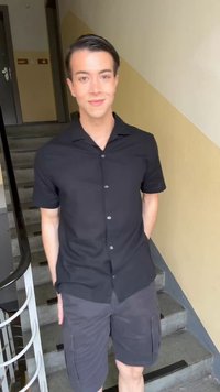 Νεαρό άτομο που στέκεται σε εσωτερικό χώρο σε σκάλα, φοράει μαύρο κοντομάνικο πουκάμισο με κουμπιά και γκρι βερμούδα cargo, χαμογελάει στην κάμερα.