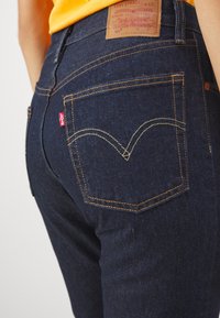Jeans in denim blu scuro con cuciture gialle, caratterizzati da un design classico della tasca posteriore e un'etichetta in pelle sulla cintura che mostra il marchio.