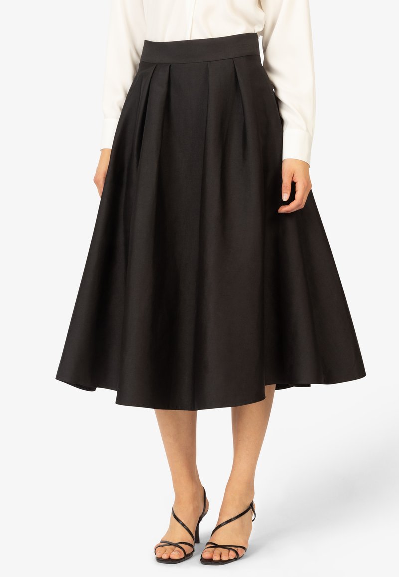 Jupe plissée noire avec une large taille, un tissu fluide et une silhouette en A, associée à des talons fins noirs.