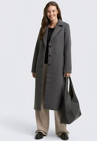 Manteau en mélange de laine gris avec une silhouette classique, col à revers et quatre boutons. Associé à un pantalon beige et un sac à main noir.