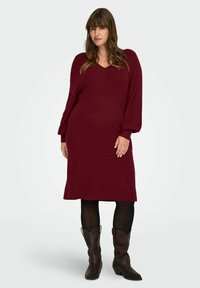 Donna che indossa un vestito a maglia bordeaux a maniche lunghe, collant neri e stivali da cowboy marroni, in piedi contro uno sfondo chiaro e semplice.