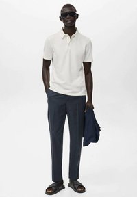 White polo shirt, dark trousers, black sandals, holding a navy jacket. Sunglasses worn. Smooth fabric, tailored fit, minimalistic design.
