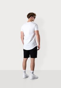Weißes T-Shirt mit kurzen Ärmeln und abgerundetem Saum, kombiniert mit schwarzen Shorts. Weiße Sneaker und Crew-Socken sichtbar; keine Logos oder Muster.