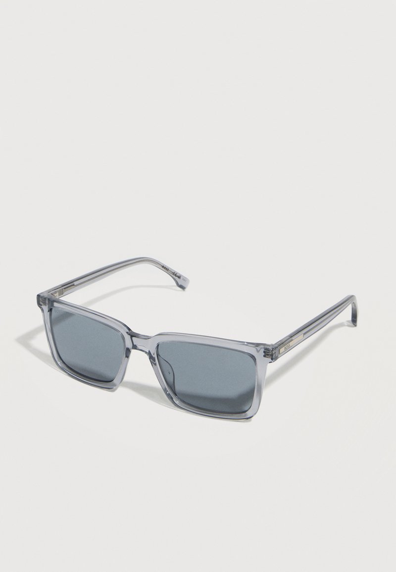 Ορθογώνια γυαλιά ηλίου με γκρι απόχρωση, διάφανα πλαστικά γκρι σκελετά και λεπτούς βραχίονες, παρουσιασμένα σε λευκή επιφάνεια.