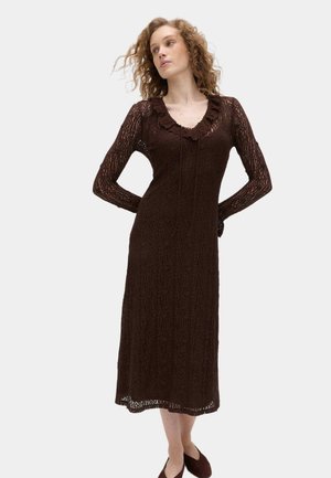 Kvinde med krøllet hår iført en brun, tekstureret midi-kjole med lange ærmer og flæsekrave, stående med hænderne bag ryggen.