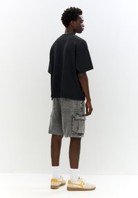 T-shirt noir oversize, shorts cargo gris avec poches, chaussettes blanches et baskets beiges avec accents jaunes. Couleurs unies et coupe décontractée.