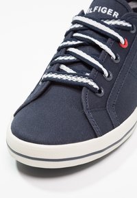 Baskets bleu marine avec des lacets rayés blancs et bleu marine, semelle en caoutchouc blanche avec une bande sombre, et un œillet rouge près du sommet.