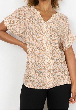 Femme portant un chemisier à manches courtes avec des rayures ondulées beige, orange et marron, associé à un pantalon noir sur un fond uni.