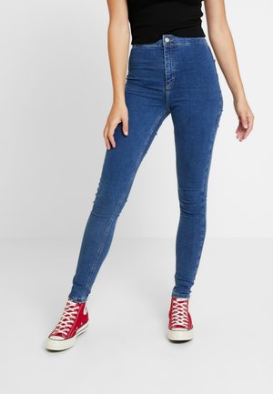 Kobieta ubrana w niebieskie dopasowane jeansy z wysokim stanem i czerwone wysokie trampki, stojąca z jedną nogą lekko wysuniętą do przodu na jednolitym tle.