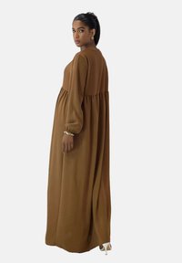 Braunes Maxikleid mit langen Ärmeln, plissierter Taille und weicher Textur. Es hat einen hohen Ausschnitt und wird mit weißen Absatzschuhen kombiniert.