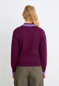 Lila kötött pulóver, amely lila és piros csíkos gallérral, bordázott aljjal és hosszú ujjakkal rendelkezik, olíva zöld cargo nadrággal viselve.