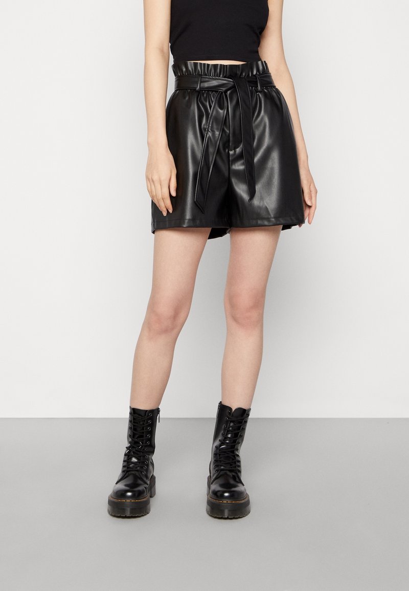 Černé šortky z umělé kůže s vysokým pasem a zavazovacím páskem, s hladkou texturou a volnějším střihem, doplněné černými kotníkovými botami na šněrování.