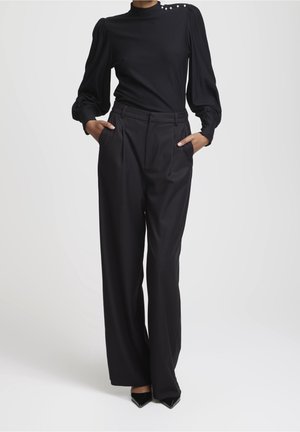 Femme portant un chemisier noir à col montant avec des manches bouffantes et des boutons en perle, associé à un pantalon noir large taille haute et des talons noirs.