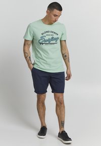 Světle mátově zelené tričko s grafikou "Blend Denim Surfing", zkombinované s námořnickými modrými šortkami. Černé tenisky dokončují tento ležérní outfit.